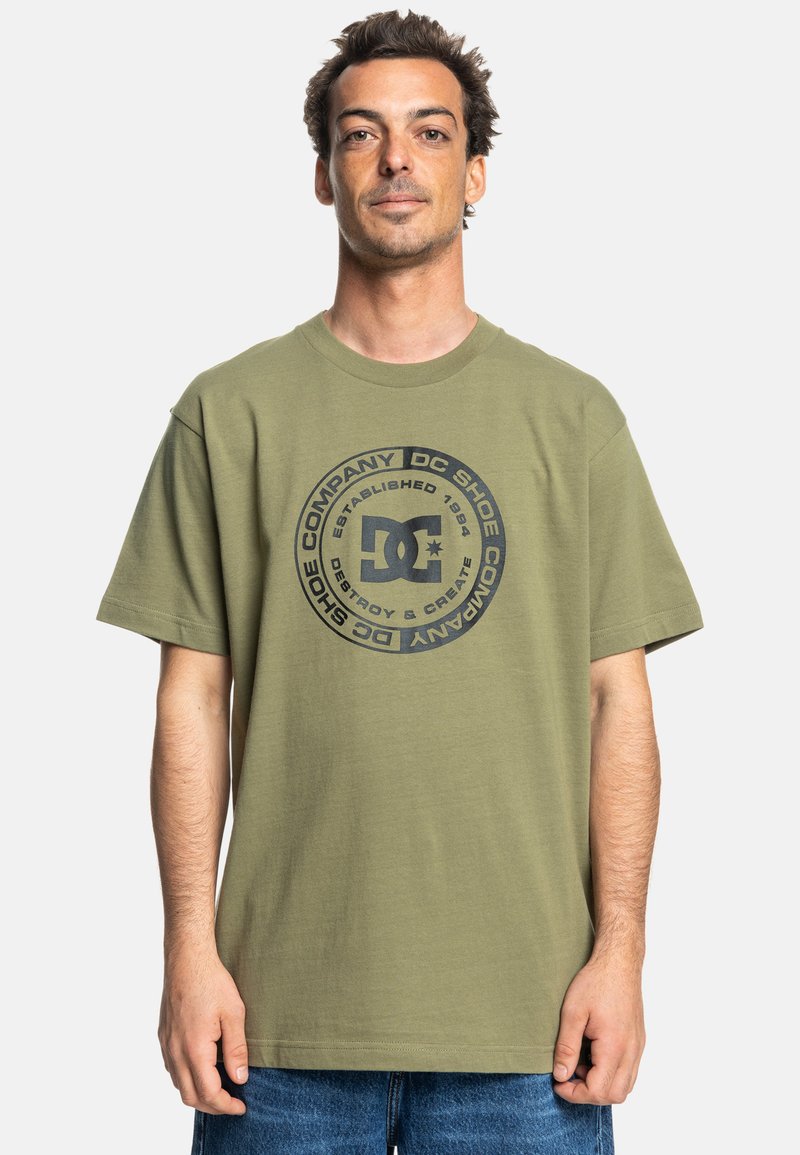 Olivegrüne Baumwoll-T-Shirt mit schwarzem runden Logo und dem Text "DC Shoe Company" sowie dem Gründungsjahr. Kurze Ärmel und entspannter Schnitt.