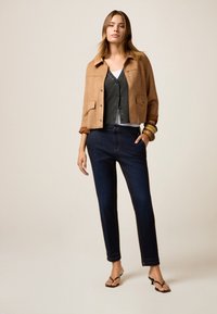 Giacca cropped in suede beige con bottoni a scatto sopra un cardigan grigio e una t-shirt bianca, abbinata a jeans scuri a sigaretta e ciabatte nere.