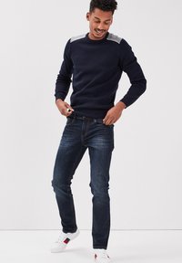 Marine gebreide trui met grijze textuuraccenten op de schouders, slim-fit donkerdenim jeans en witte sneakers met gekleurde streepdetails.