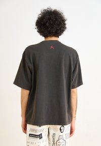Camiseta negra de gran tamaño hecha de algodón, con un escote redondo y mangas cortas. Un logo rojo de Jordan se encuentra en la parte posterior cerca del escote.
