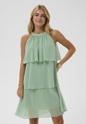 Jeune femme portant une robe verte menthe sans manches à plusieurs couches avec un décolleté dos nu, un bracelet en or et des boucles d'oreilles créoles, se tenant devant un fond uni.