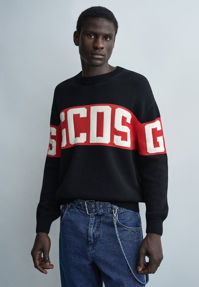 Homme portant un pull noir avec des lettres rouges et blanches sur la poitrine et les manches, associé à un jean bleu et une ceinture à œillets métalliques.