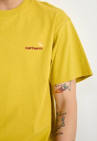 Geel katoenen t-shirt met een ronde hals, korte mouwen en een klein geborduurd Carhartt-logo in oranje en bordeaux.