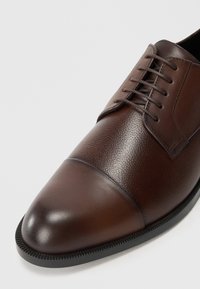 Brown leather dress shoe with cap toe, laces, and textured upper, on a light background.