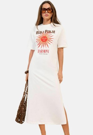 Kobieta nosząca białą sukienkę midi z motywem słońca i napisem „BALI PALM HARMONY”, duże brązowe okulary przeciwsłoneczne, drewniana bransoletka i pleciona torba.