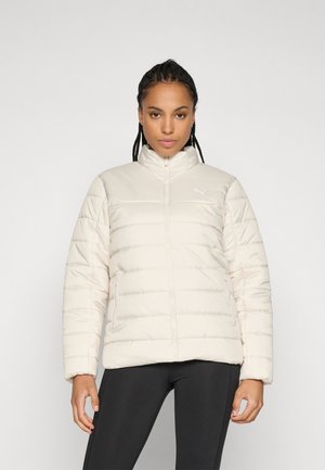 Femme avec des cheveux tressés portant une veste matelassée beige clair à fermeture éclair et un legging noir, se tenant devant un fond gris clair uni.