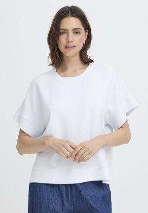 Fiatal nő vállig érő barna hajjal, laza, rövid ujjú fehér felsőt és kék nadrágot visel, egyszerű háttér előtt áll.