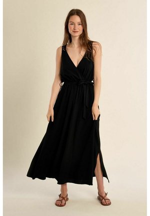 Donna in piedi con un abito nero avvolgente senza maniche, con spacco laterale e cintura, indossando sandali piatti marroni su uno sfondo neutro.