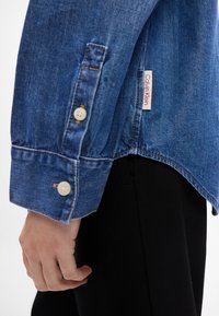 Camicia in denim con polsini abbottonati, colore azzurro chiaro, e etichetta bianca con "Calvin Klein". Cuciture visibili e texture liscia.