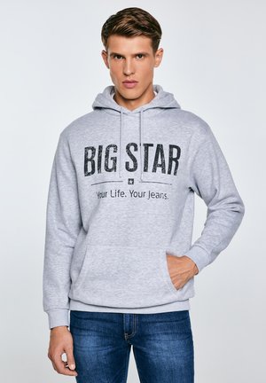 Mladý muž v světle šedé mikině s kapucí s nápisem "BIG STAR Your Life. Your Jeans." a modrých džínách stojí před jednobarevným pozadím.