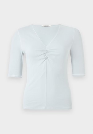 Femme portant un haut bleu clair froncé, une jupe midi noire à imprimé floral vert, et des sandales noires à talons hauts à brides, debout sur fond blanc.