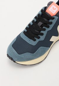 Marineblå og blå sneakers med semsket skinn og teksturert stoff, svarte snøringer, beige såle og rosa logo-merke på tungen. Rund tådesign.