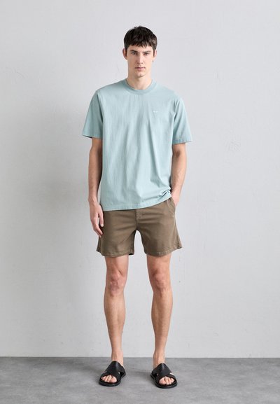 Lys blå bomulls t-skjorte med rund hals, kombinert med brune shorts og svarte sandaler. Modellen står mot en nøytral bakgrunn.