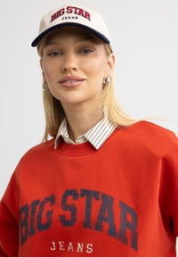 Punane pusakas, millel on tumevetru kirjaga "BIG STAR JEANS", on kihitatud valge ja musta triibulise kraega särgi peale. Beez müts sarnase kirjaga.