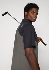 Πόλο γκολφ κοντομάνικο, μαύρη μπλούζα με γκρι κάτω τμήμα, απαλό υφή, διαθέτει γιακά και λεπτομέρεια λογότυπου στον ώμο, με κρατημένο το γκολφ κλαμπ.