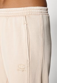 Ljusbeige mjukisbyxor i mjukt material, med elastisk midja, sidofickor och en broderad logotyp på vänster lår.