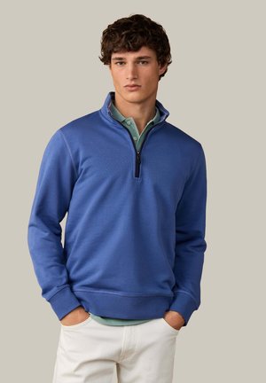 Hombre joven con un jersey azul con cremallera hasta la mitad, sobre una camisa polo verde y pantalones blancos, con las manos en los bolsillos, sobre un fondo liso.