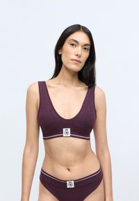 Violetti puuvillainen bralette, jossa on leveät hihnat ja syvä V-pääntie. Siinä on logopatchi ja koristeellinen raidallinen nauha alaosassa.