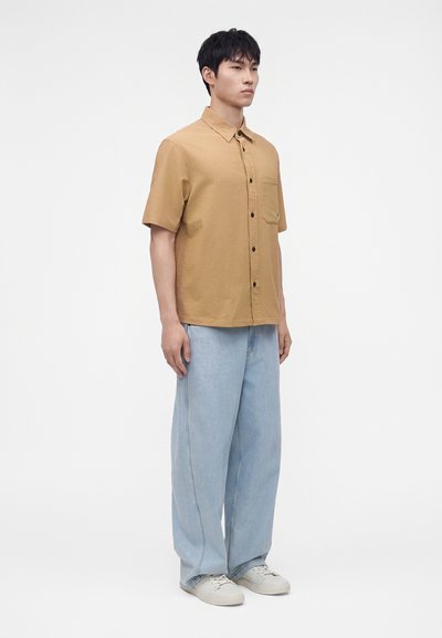 Homme debout portant une chemise beige à manches courtes avec des boutons, un jean large bleu clair et des baskets blanches sur fond blanc.
