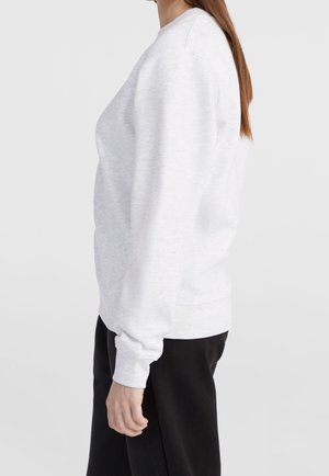 Vrouw die een effen lichtgrijze trui en zwarte broek draagt, zijaanzicht tegen een effen witte achtergrond.