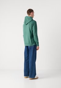 Felpa verde con cappuccio, dalla texture liscia, abbinata a jeans di denim blu larghi e sneakers bianche, vista da dietro.