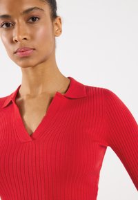 Rotes geripptes Strickoberteil mit V-Ausschnitt und kurzen Ärmeln. Ausgestattet mit einfarbigem Design und einem Kragen, der die figurschmeichelnde Passform betont.