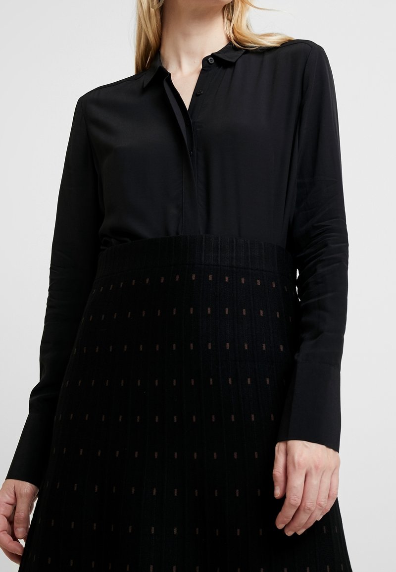 Blusa nera a maniche lunghe con colletto a punta, abbinata a una gonna nera strutturata con strisce verticali e piccoli quadrati marroni.