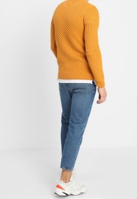 Personne portant un pull moutarde texturé, un jean bleu, des baskets blanches avec des accents orange, debout devant un fond blanc uni.