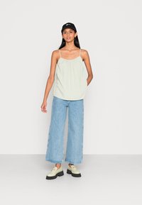 Top sem mangas verde-claro com decote franzido, combinado com calças de ganga largas azul-claro e sapatos pretos com sola grossa.