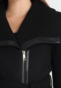 Cappotto di lana nero con un ampio colletto, caratterizzato da un accento in pelle e una zip metallica centrale. Include una vita con cintura per una vestibilità regolabile.