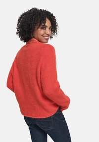 Maglione rosso, pelo soffice, con scollatura rotonda e spalle scese, abbinato a jeans blu scuro. Dettaglio discreto sul retro vicino al collo.