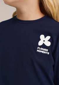 Tamsiai mėlynas medvilninis marškinėliai ilgomis rankovėmis su baltu gėlės logotipu ir užrašu "GĖLIŲ MOMENTAI" kairėje krūtinės pusėje. Paprasto apvalios kaklo dizainas.
