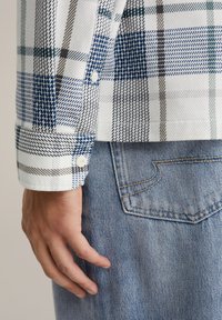 Valkoinen ja sinivalkoruutuinen paita, jossa napilliset kalvosimet, yhdistetty vaaleansinisiin farkkuihin, joissa takatasku ja näkyvät ompeluohjeet.