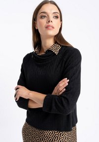 Czarny sweter o teksturze z golfem, nałożony na beżową koszulę z kołnierzykiem w geometryczne wzory z zapięciem na guziki.