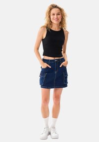 Denimrok met twee grote voorkruiven, halverwege de dij en in de kleur blauw; gecombineerd met een zwart mouwloos topje en witte sneakers.