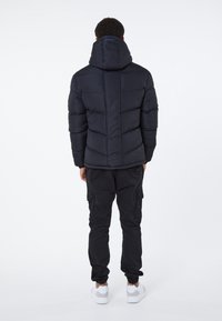 Uomo che indossa una giacca a vento nera con cappuccio imbottito, pantaloni cargo neri e sneakers bianche, in piedi rivolto verso l'esterno di fronte a uno sfondo bianco uniforme.