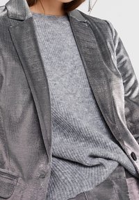 Stříbrný metalický blazer se strukturovanými klopami, kombinovaný s měkkým šedým žebrovaným svetrem. Blazer má lesklou texturu a přiléhavý střih.