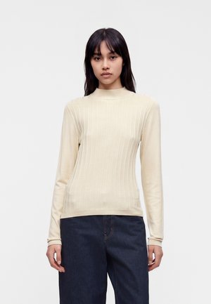 Ung kvinde med glat mørkt hår iført en cremefarvet ribbet langærmet sweater og mørkeblå jeans, stående mod en ensfarvet lys baggrund.