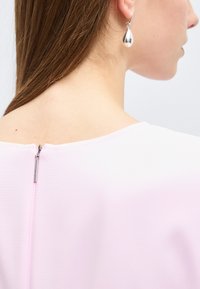 Femme aux longs cheveux bruns portant des boucles d'oreilles pendantes en argent et un haut rose pâle avec une fermeture éclair au dos sous le col.