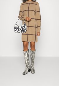 Una persona con i capelli ricci indossa un vestito a maglione a scacchi beige, tiene una borsa a tracolla con un motivo in bianco e nero e indossa stivali a righe fino al ginocchio.