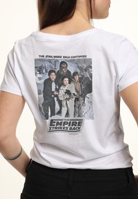 Hvid bomulds t-shirt med et grafik af fire karakterer fra "The Empire Strikes Back," med tekst over og under billedet.