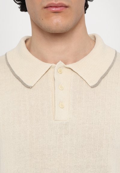 Beige színű pólóing texturált kötött anyagból, szürke színű gallérral és három gombos lezárással. Puha anyag, szűkített fazonnal.