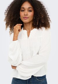 Γυναίκα με σγουρά μαλλιά που φοράει λευκό μπλουζάκι με μακριά μανίκια και υφή, με λεπτομέρειες δαντέλας, και μπλε τζιν, ποζάροντας με το ένα χέρι κοντά στο πρόσωπό της.