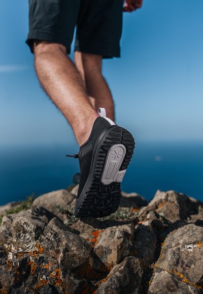 Black lightweight sneaker with textured rubber sole, elastic lacing, and pull tab; positioned on rocky terrain against a blue sky background.