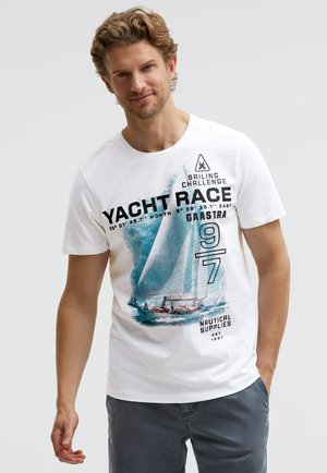 Man wearing white t-shirt with blue sailboat graphic and text "Yacht Race," standing with one hand in pocket against plain background.
