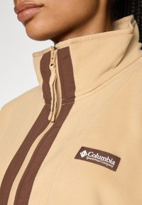 Béžová fleecová bunda s lemeom na zips, hnedými pruhmi a nášivkou loga Columbia Sportswear na hrudi. Mäkká textúra.