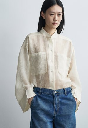 Νέο άτομο με ίσια μαύρα μαλλιά, φορώντας διαφανές μπεζ πουκάμισο με μεγάλες τσέπες και μπλε τζιν, με τα χέρια στις τσέπες.