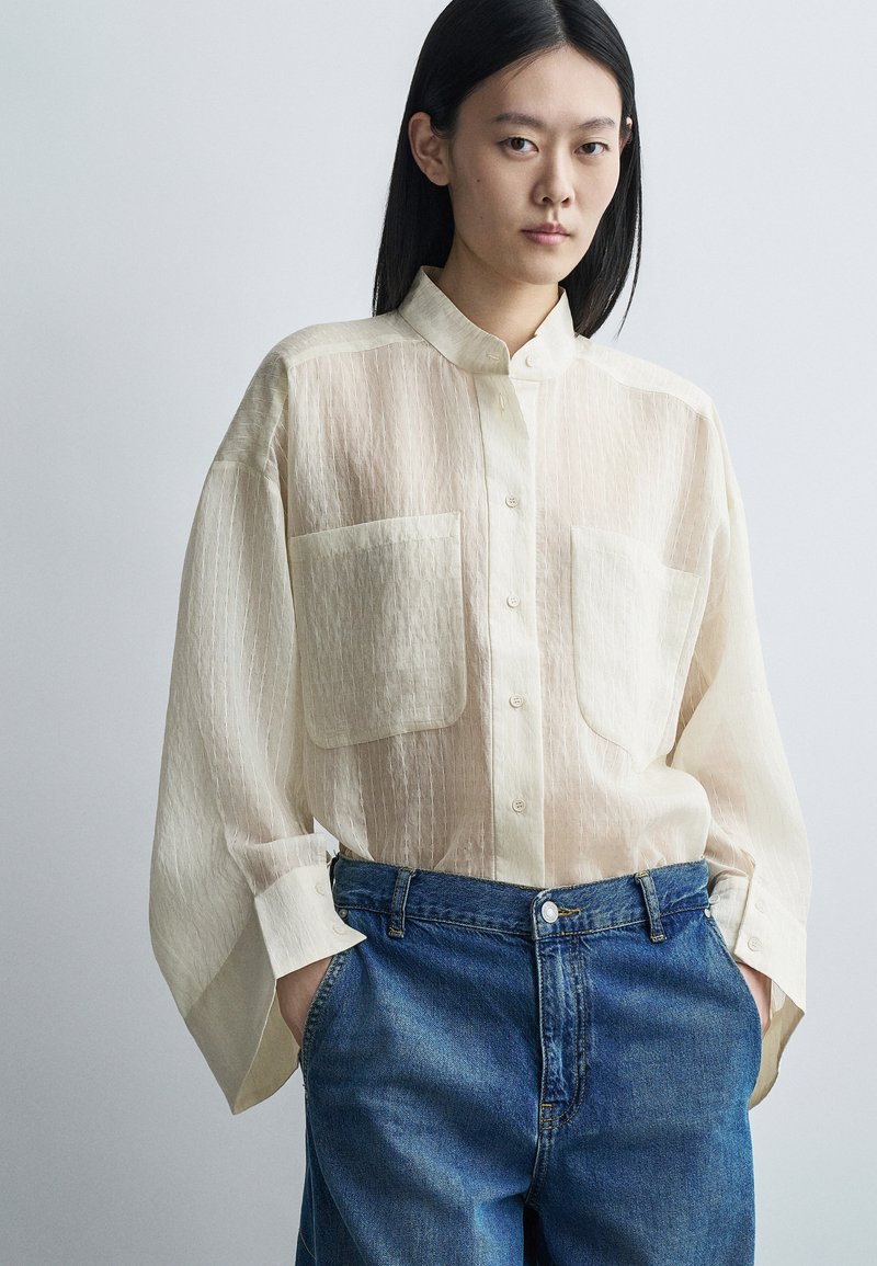 Jeune personne aux cheveux noirs raides portant une chemise beige transparente boutonnée avec de grandes poches et un jean en denim bleu, mains dans les poches.