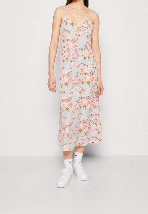 Nainen pukeutunut vaaleansiniseen midimekkoon, jossa on vaaleanpunaisia kukkakuvioita, valkoisiin Nike-sukkiin ja valkoisiin tennareihin, seisoo yksivärisen taustan edessä.