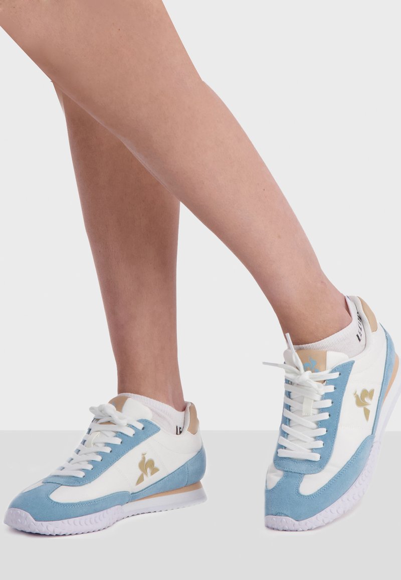 Sneaker blu e bianchi realizzati in materiali scamosciati e in pelle, con un logo dorato, lacci bianchi e una suola bianca texturizzata.